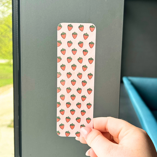 Hand holding a pink strawberry bookmark in front of a soft grey background and window view.